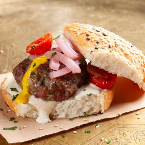 Panecillo artesano de la casa con mini hamburguesa de kebab ahumado con tahina, salsa amba de mango, vinagre y especias, tomate asado y cebolla envinagrada.