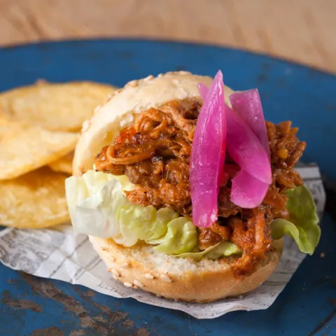 "Minihamburguesa de carne de cerdo desmenuzada, ahumada a baja temperatura, con cebolla envinagrada casera y chiles de Jalapa mexicanos, acompañado con patatas gruesas."