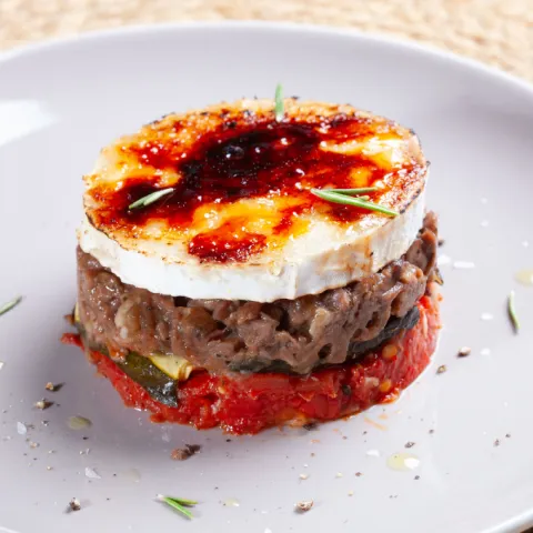 Timbal crujiente de patatas, calabacín, mejilla de cerdo ibérico a baja temperatura, tomates encurtidos y queso de cabra gratinado, con un toque de miel de caña.