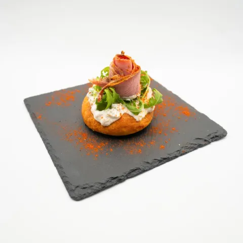 Sobre pan de salaílla, una base de tzatziki de pepinillos, una cama de rúcula y finas lonchas de pastrami en forma de flor,coronada con unas gotitas de AOVE y pimentón De la Vera
