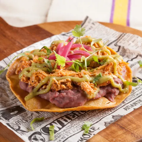 Tostada de maíz cubierta con frijoles refritos y tinga de pollo, con mayonesa de chipotle, cilantro y cebolla encurtida.