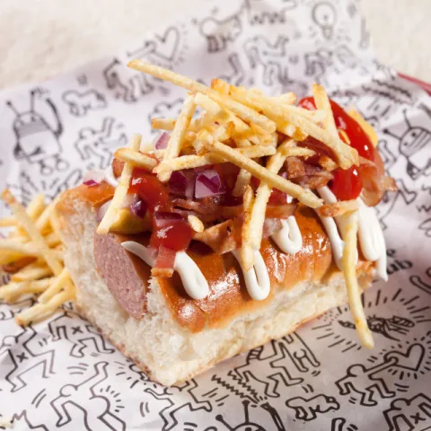 Pan artesano con Frankfurt, mostaza, mayonesa cremosa, cebolla cortada en brunoise, bacon crujiente y patatas paja.