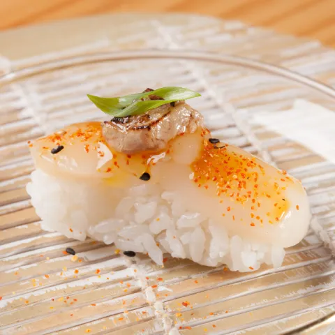 Base de arroz de sushi cubierta con una porción de vieira flambeada, salsa unagi, foie-gras, shichimi, cebollino y sal Maldon.