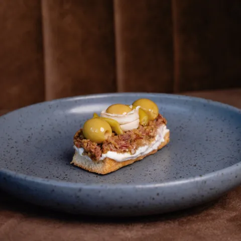 Tosta de jamón ibérico deshilachado y aliñado con mostaza antigua, coronada con aceitunas rellenas, piparra y boquerón
