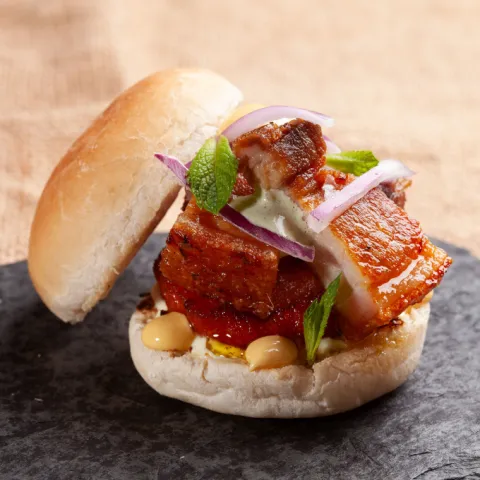 Pan con mayonesa ahumada, rodajas de boniato, chicharrones de cerdo, menta, limón, cebolla morada, salsa criolla y gotas de huancaína.