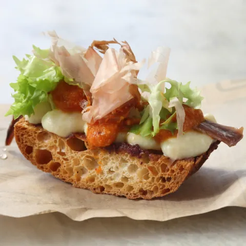 Tostada crujiente con manteca de aceitunas de Kalamata, crema de bacalao, anchoa, nuestra salsa xatonaire, láminas de katsuobushi y escarola