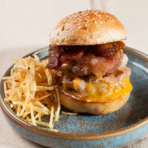 Jugosa hamburguesa de carne de ternera, queso cheddar fundido y mermelada de bacon sobre una base crujiente de patatas paja.