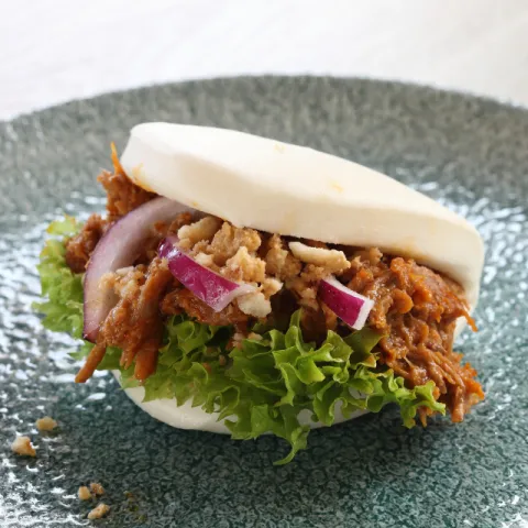 Bocadito de pan bao relleno de melosa carrillera, curry rojo y coco, torreznos y cebolla encurtida